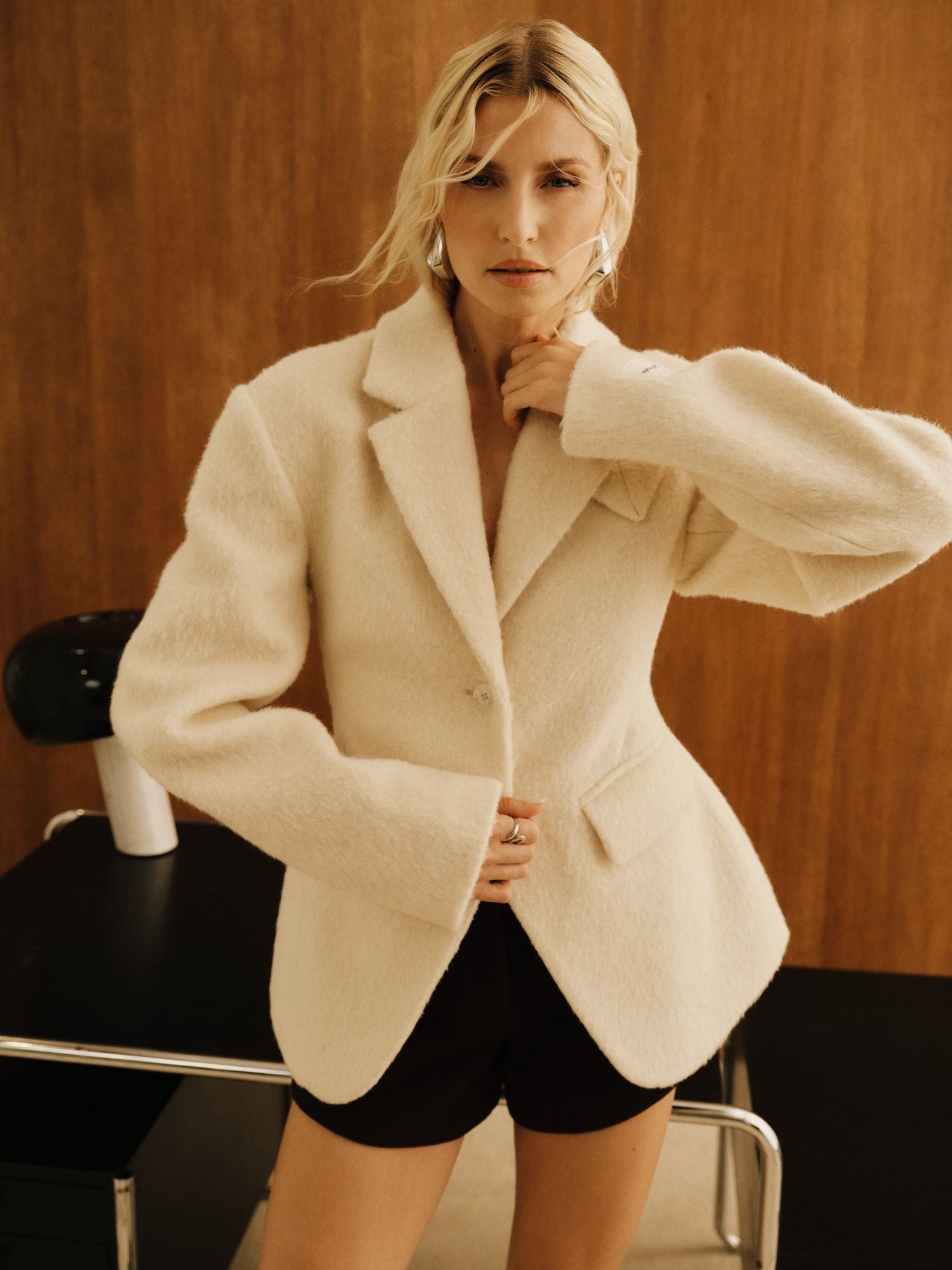 Eine Frau mit welligem blondem Haar trägt die Jacke „Mila“ von LeGer by Lena Gercke – eine cremefarbene, strukturierte Damenjacke –, dazu schwarze Shorts, während sie in einem Innenraum vor einer Holzwand steht und mit einer Hand ihren Kragen zurechtzieht. Hinter ihr steht eine schwarz-weiße Lampe auf einem Schreibtisch.