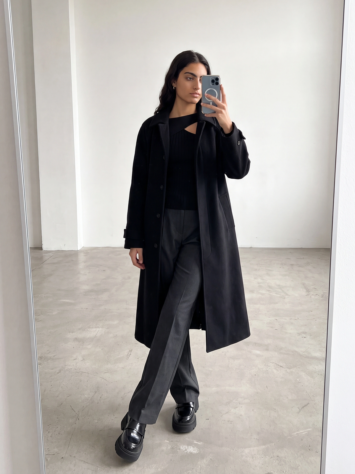 Eine Frau macht in einem minimalistisch eingerichteten Raum ein Spiegel-Selfie. Sie trägt den Mantel „Sybille“ von LeGer by Lena Gercke mit einer eleganten einreihigen Knopfleiste, dazu ein schwarzes Oberteil, eine dunkelgraue Hose und schwarze Schuhe. Sie steht mit überkreuzten Beinen da und hält ihr Handy mit der rechten Hand in der Hand.