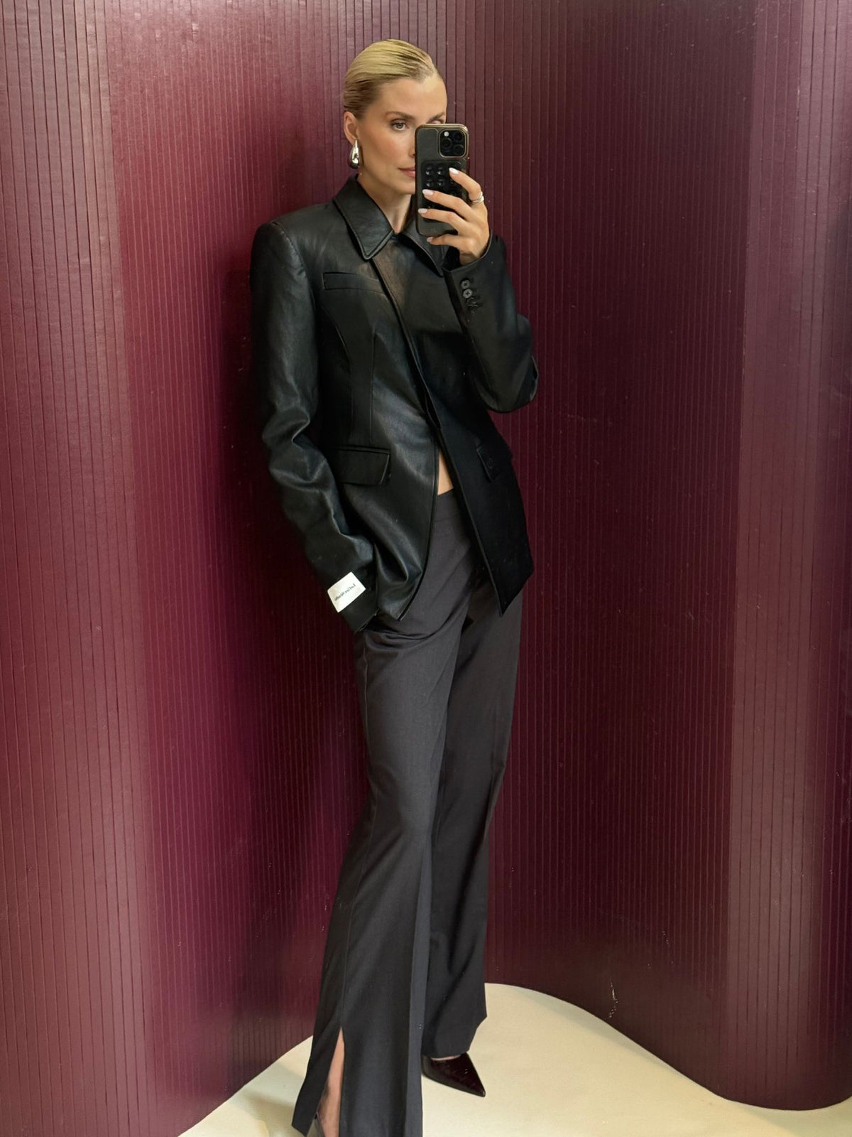 Eine Person steht vor einer kastanienbraunen Wand, trägt einen schwarzen Lederblazer und die Hose „Fergie“ von LeGer by Lena Gercke – eine dunkle Hose mit weitem Bein und Schlitzen am Saum in der Passform „Tall“ – sowie Ohrringe und macht mit ihrem Handy ein Spiegel-Selfie.