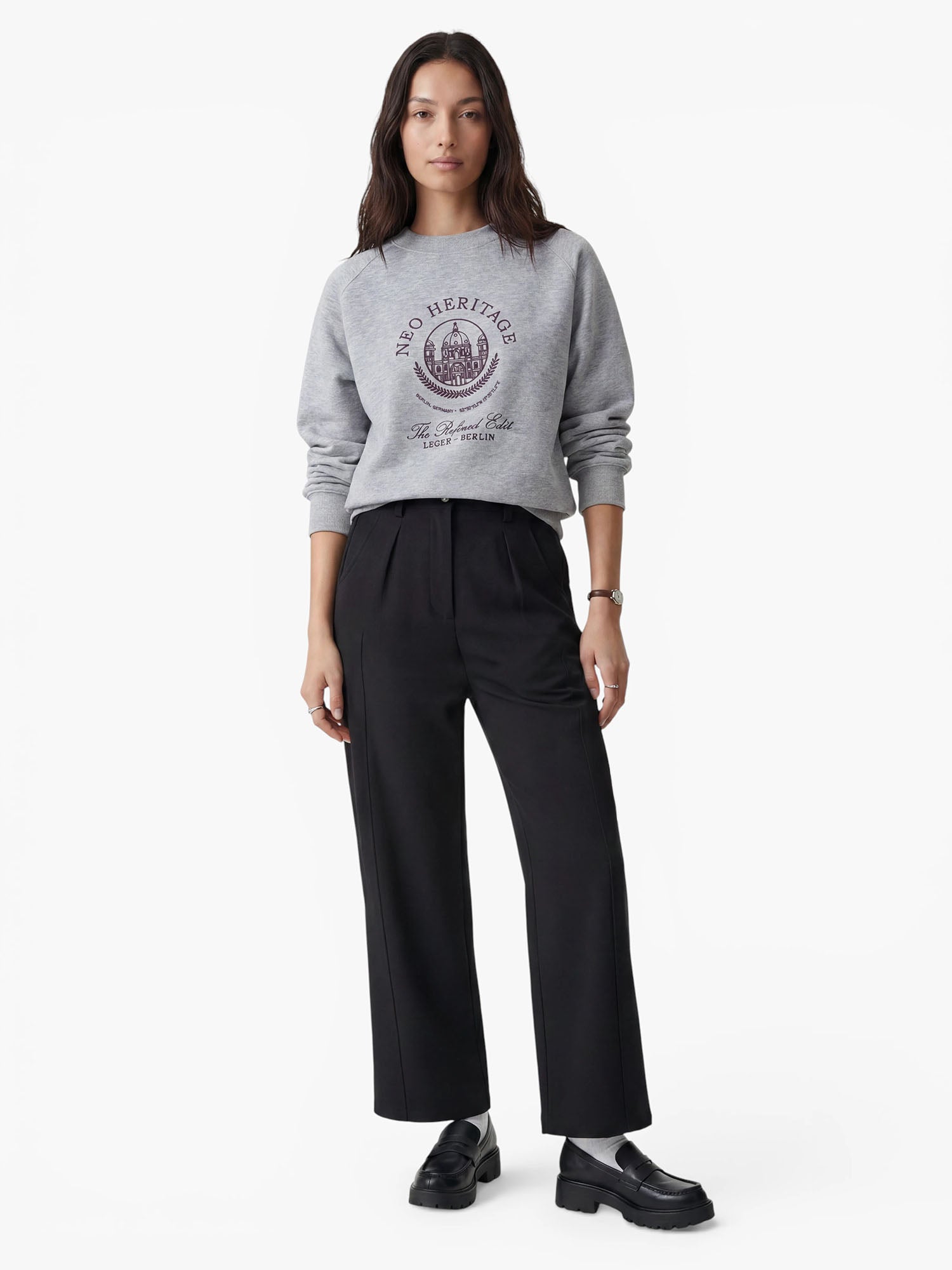 Vor einem schlichten weißen Hintergrund steht eine Frau mit langen Haaren. Sie trägt ein graues Sweatshirt mit Aufdruck und Emblem, dazu eine schwarze Plisseehose „Kasha“ von LeGer by Lena Gercke und schwarze Loafer.