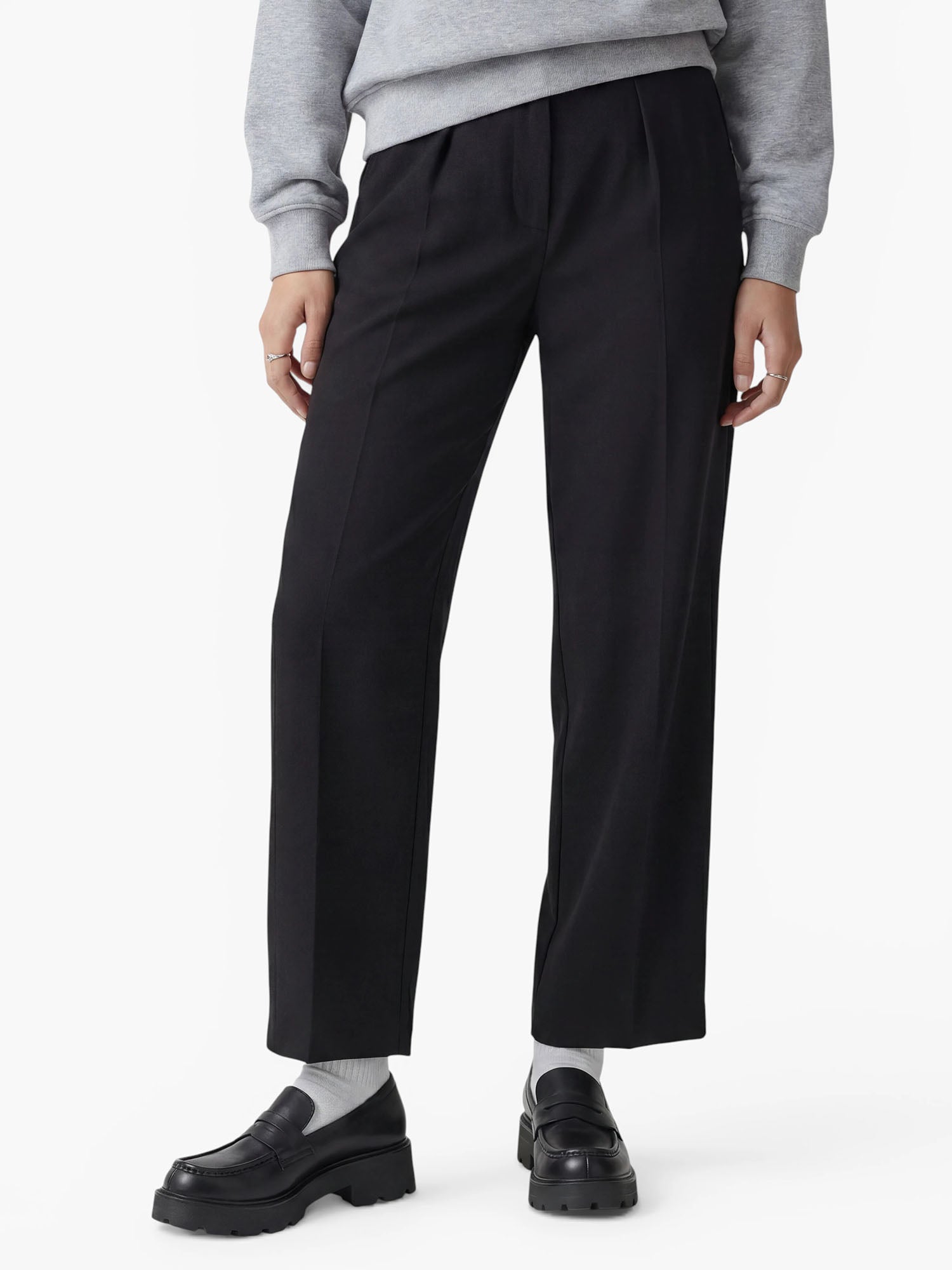 Eine Person, die die Hose „Kasha“ von LeGer by Lena Gercke in schwarzer Ausführung mit weitem Bein trägt und diese mit schwarzen Loafern, weißen Socken und einem hellgrauen Pullover kombiniert hat, steht vor einem schlichten weißen Hintergrund. Nur die untere Körperhälfte ist zu sehen.
