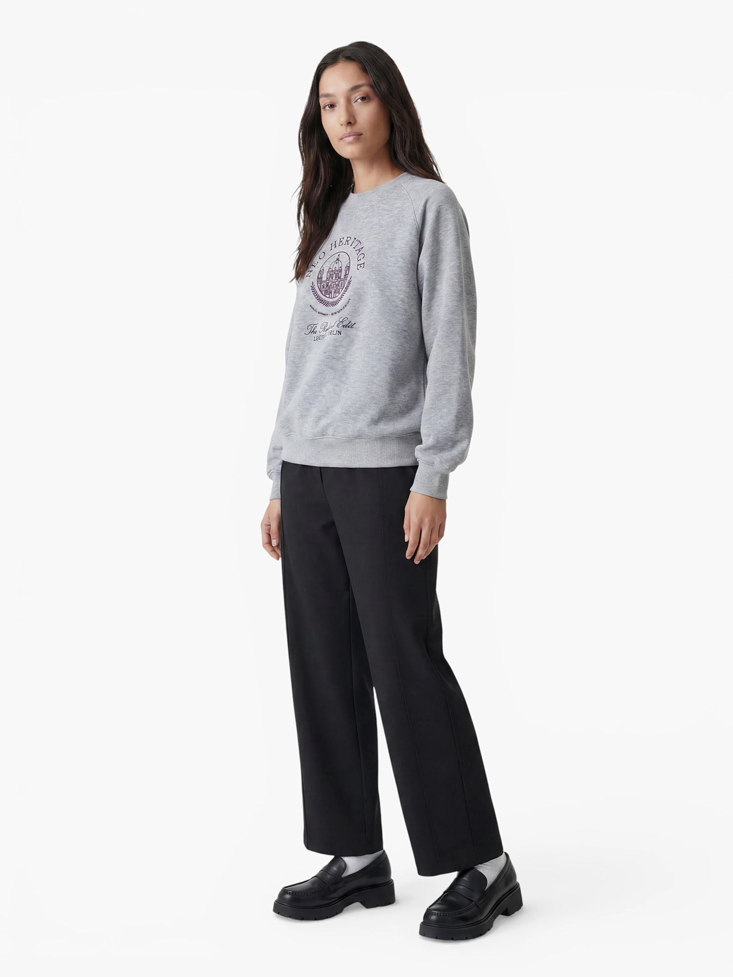 Eine Person steht vor einem weißen Hintergrund und trägt ein graues Sweatshirt mit Aufdruck, eine schwarze, taillierte „Kasha“-Hose von LeGer by Lena Gercke sowie schwarze Loafer. Sie hat langes dunkles Haar und einen neutralen Gesichtsausdruck.