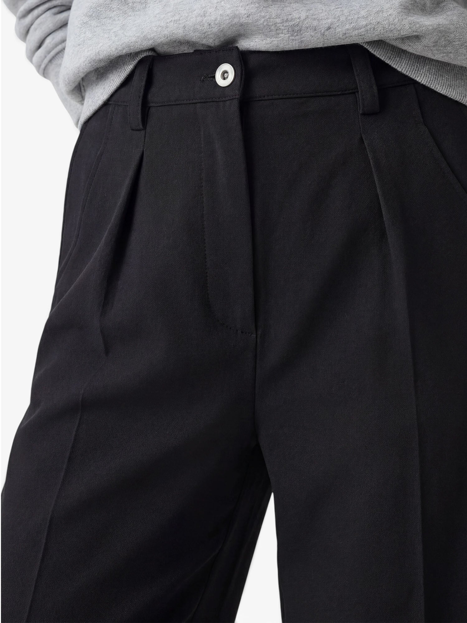Nahaufnahme einer Person, die die hoch taillierte, schwarze, taillierte Hose „Kasha“ von LeGer by Lena Gercke trägt, die sich durch Falten, Gürtelschlaufen und einen weißen Knopf auszeichnet – diese moderne Damenhose vereint Stil und Komfort. Die Person kombiniert die taillierte „Kasha“-Hose mit einem hellgrauen, in die Hose gesteckten Oberteil.