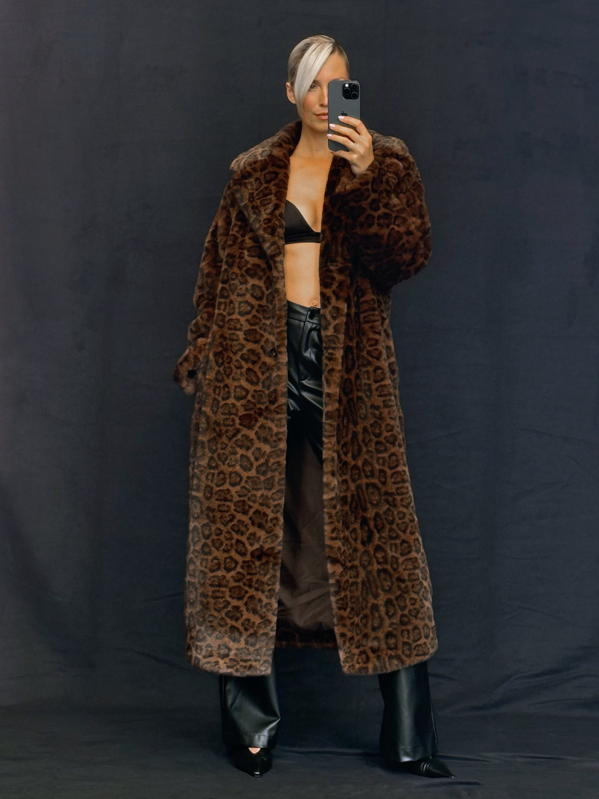 Eine Person macht ein Spiegel-Selfie in einem „Maischa“-Mantel von LeGer by Lena Gercke, einem Kunstpelzmantel mit voluminösem Kragen und Leopardenmuster, den sie über einem schwarzen BH und einer Lederhose trägt.