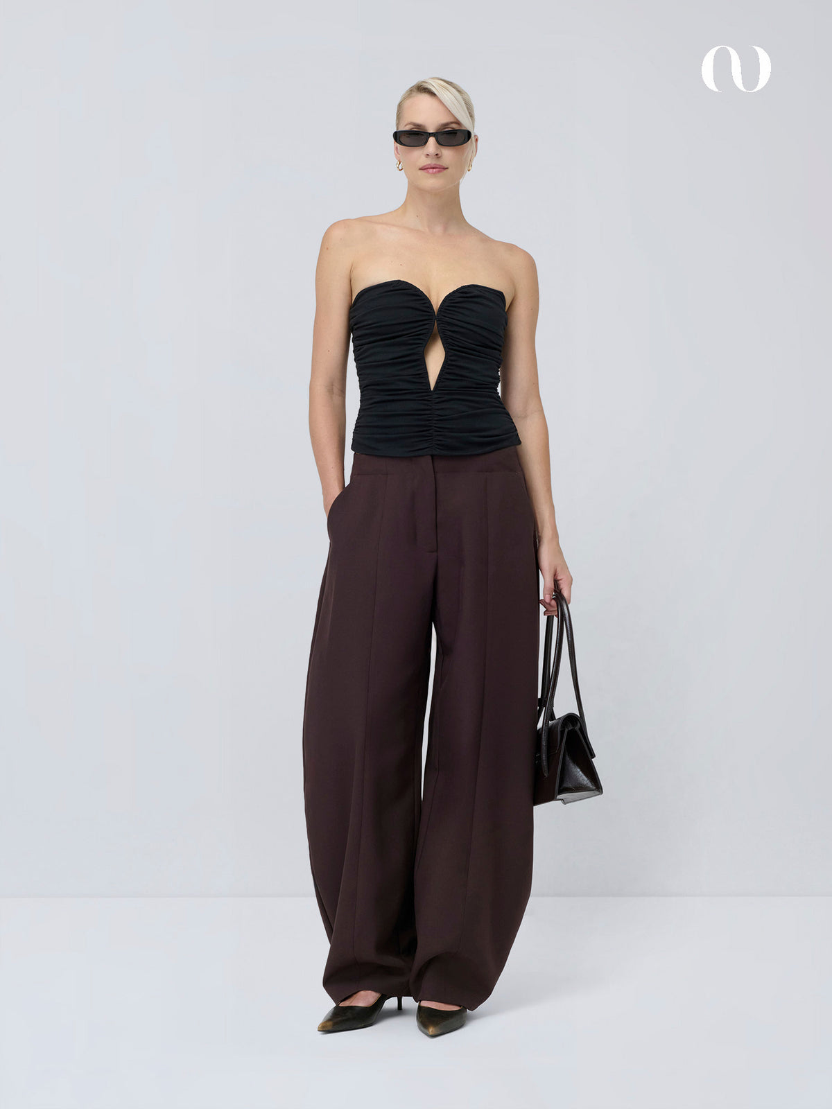 Ein Model trägt ein schwarzes „Marion“-Top im trägerlosen Design mit tiefem Ausschnitt vorne, dazu eine braune Hose mit weitem Bein und eine kleine schwarze Handtasche.