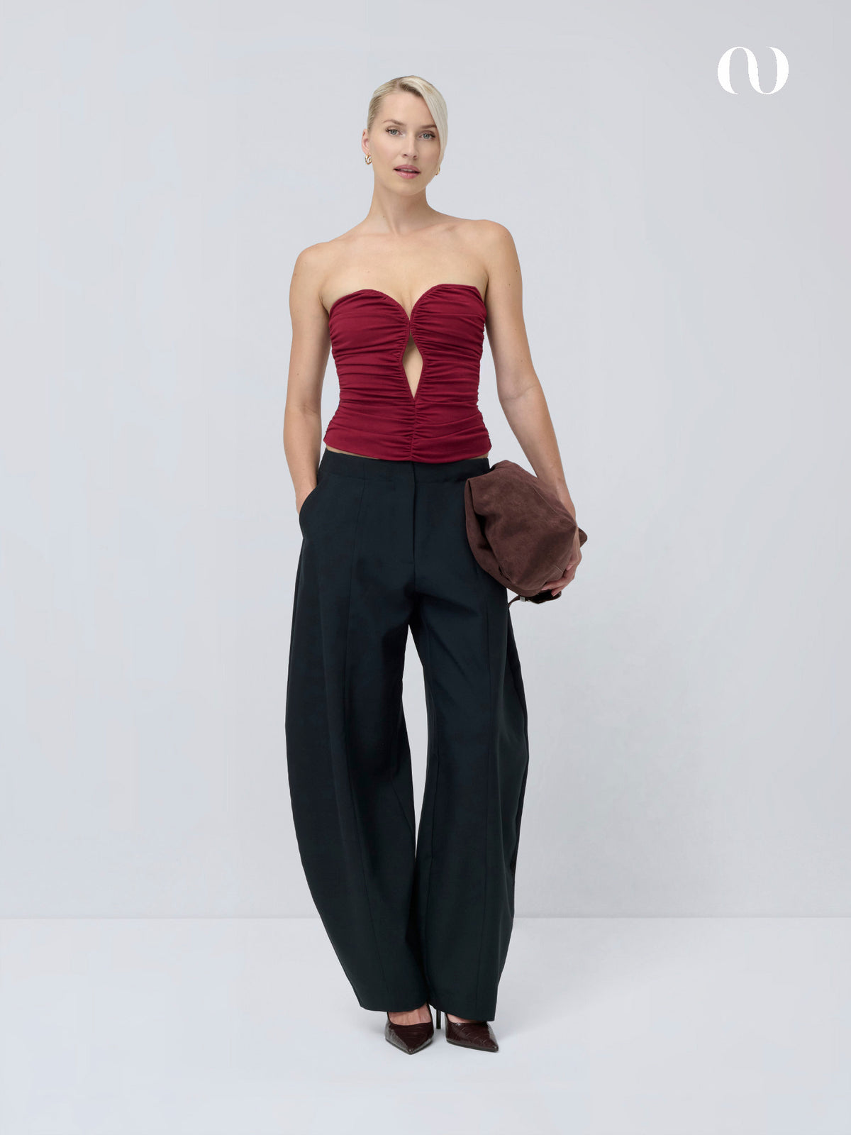 Eine Frau, die ein weinrotes „Marion“-Oberteil mit trägerlosem Herzausschnitt und gerafftem Ausschnitt vorne trägt, dazu eine schwarze Hose mit weitem Bein und eine braune Clutch.