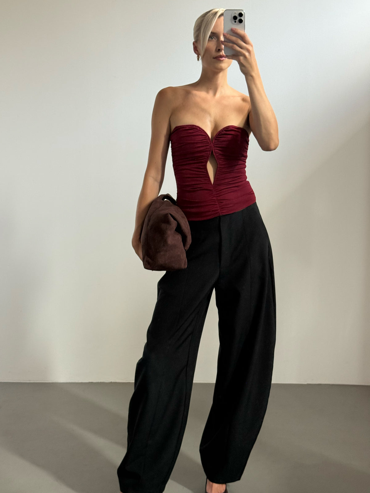 Eine Person, die das Oberteil „Marion“ trägt – ein burgunderrotes, schulterfreies, gerafftes Oberteil im Korsettstil mit Ausschnitt vorne –, kombiniert mit einer schwarzen Hose mit weitem Bein und einer großen braunen Clutch.