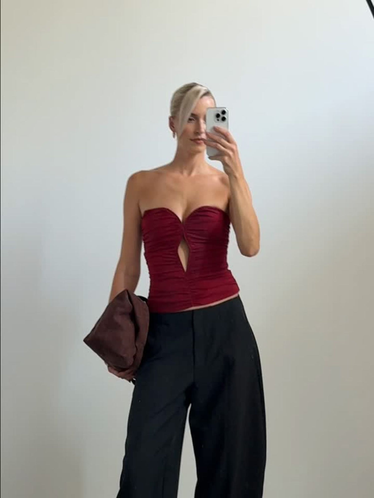 Eine Person, die das Oberteil „Marion“ von LeGer by Lena Gercke trägt – ein dunkelrotes, schulterfreies Modell mit einem Ausschnitt in der Mitte –, dazu eine schwarze Hose und eine dunkelbraune Clutch in der Hand, während sie ein Spiegel-Selfie macht.
