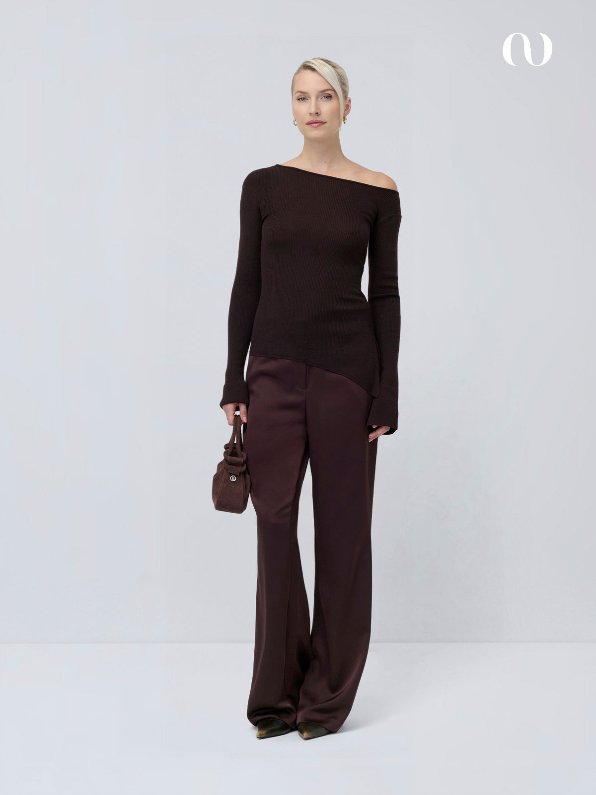 Ein Model trägt einen braunen Joelle-Pullover, eine dunkelbraune Hose und eine kleine braune Handtasche.