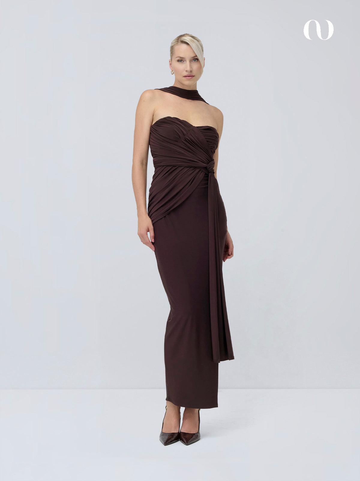 Eine Frau trägt das dunkelbraune, trägerlose, bodenlange Kleid „Thekla“ von LeGer by Lena Gercke, das sich durch eine asymmetrische Drapierung an der Taille, ein passendes Halsband im Choker-Stil und Pumps mit spitzer Zehenpartie auszeichnet.