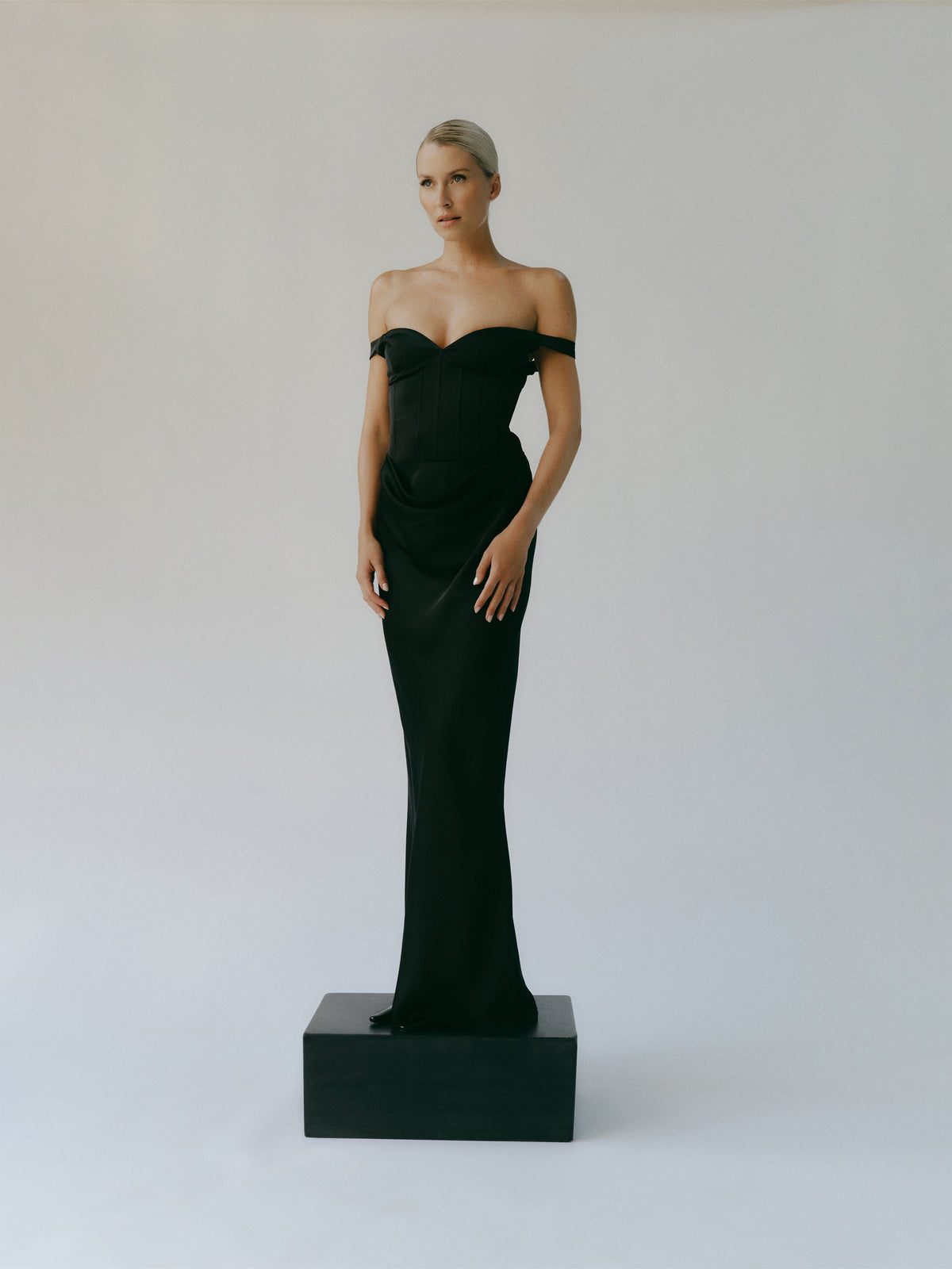 Eine Frau trägt das Kleid „Jocelyn“ von LeGer by Lena Gercke, ein bodenlanges, schwarzes, schulterfreies Abendkleid mit einem Oberteil im Korsettstil und Herzausschnitt, während sie auf einem schwarzen Block steht.