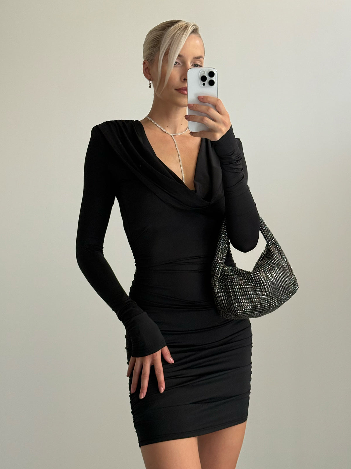 Eine Frau in einem schwarzen „Hildegard“-Kleid mit langen Ärmeln und Wasserfallausschnitt, dazu eine funkelnde schwarze Umhängetasche.