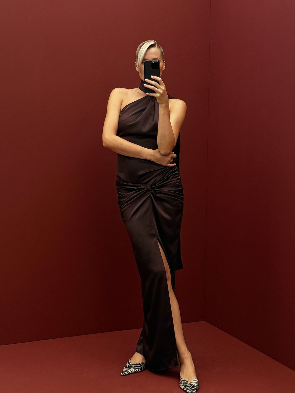 Eine Person, die ein dunkelbraunes Josephin-Midikleid mit One-Shoulder-Ausschnitt, einem Schlitz vorne und Pumps mit Zebradruck trägt und ein Spiegel-Selfie macht.