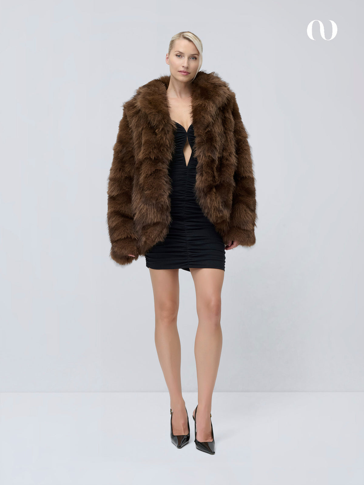 Ein Model trägt eine dunkelbraune „Dion“-Jacke aus Kunstfell über einem schwarzen Minikleid und Pumps mit spitzer Zehenpartie.