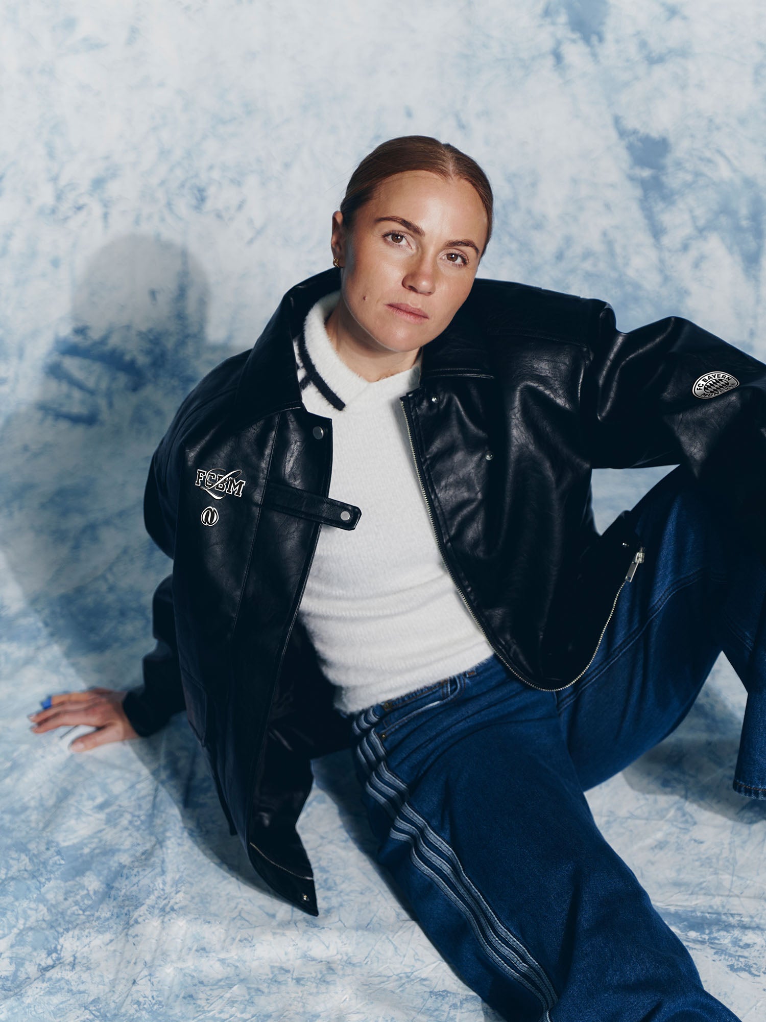 Eine hellhäutige Person mit hochgesteckten rötlichen Haaren trägt die Elisa Jacket von LeGer by Lena Gercke, einen weißen Pullover und eine blaue Jeans, während sie vor einem blau-weiß marmorierten Hintergrund auf dem Boden sitzt und selbstbewusst in die Kamera schaut.