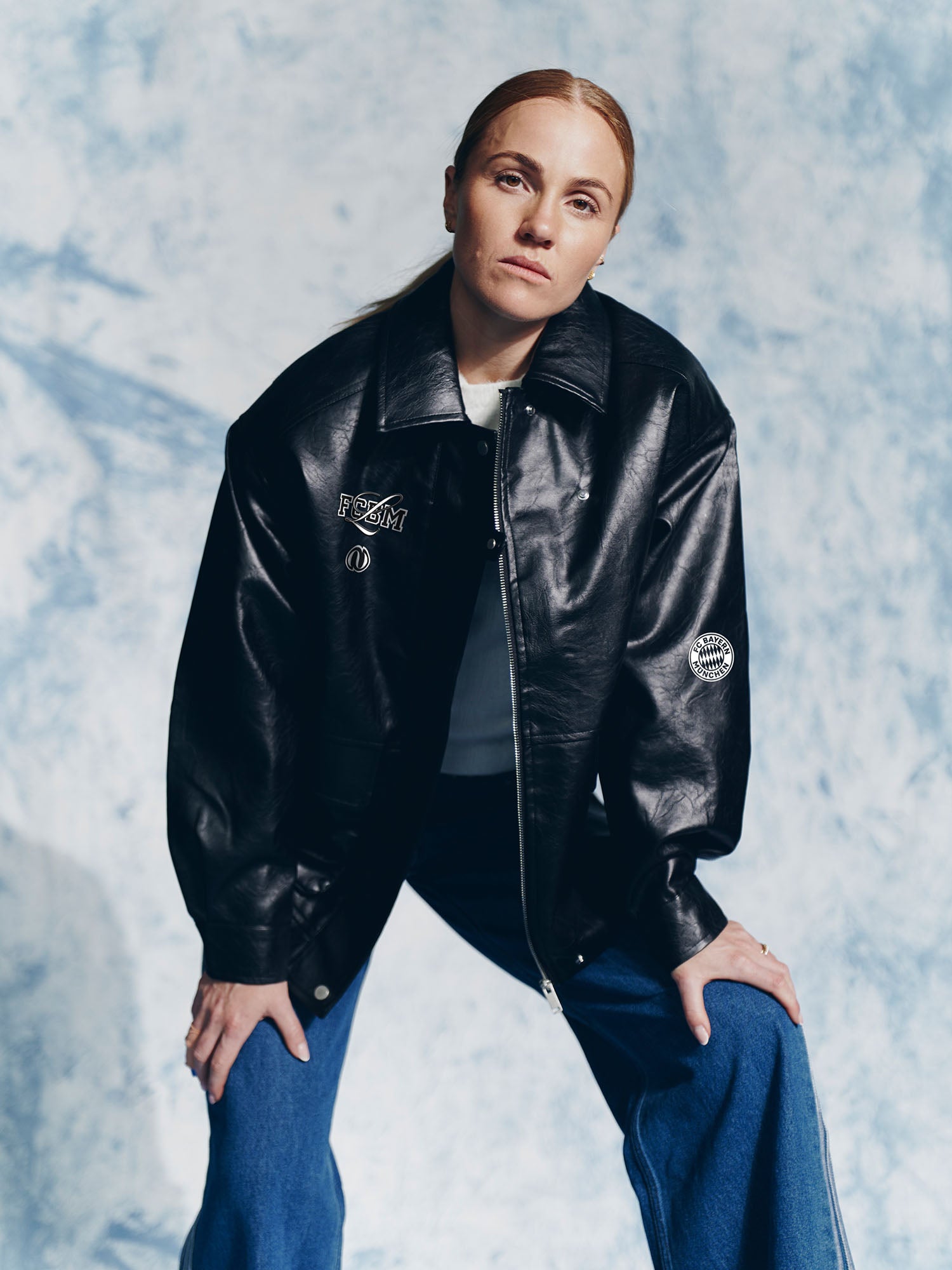 Eine hellhäutige Person mit glattem, nach hinten gebundenem rotem Haar trägt eine LeGer by Lena Gercke Elisa Jacket und blaue Jeans und lehnt sich mit den Händen auf den Knien vor einem hellblau strukturierten Hintergrund nach vorne.