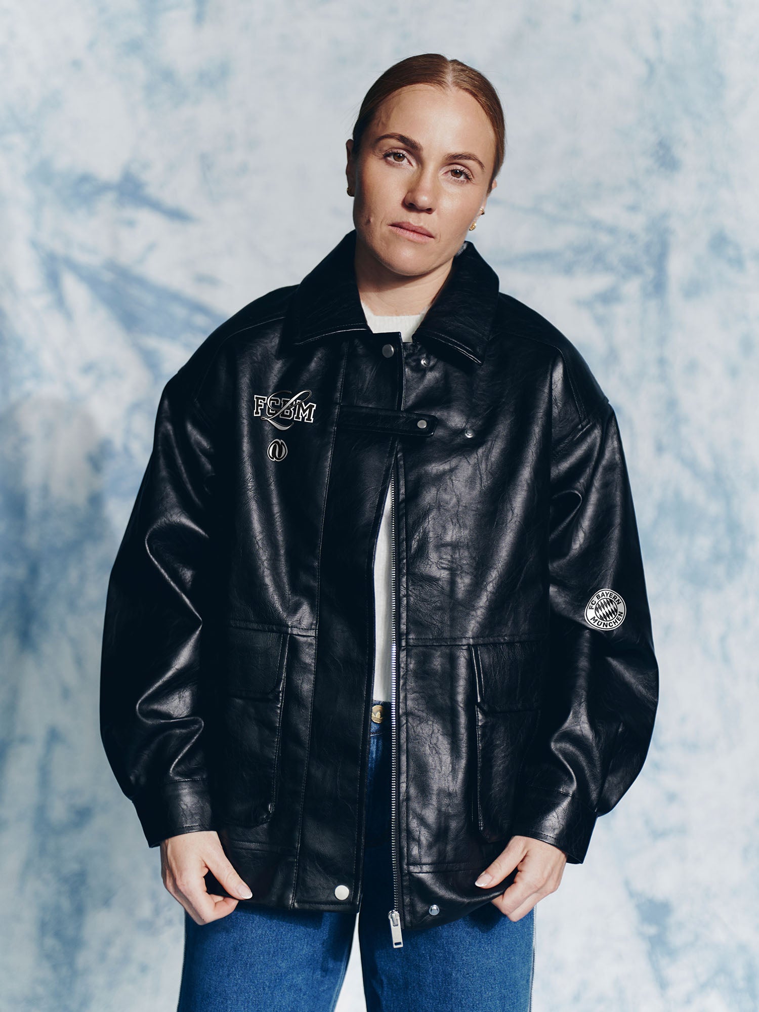 Eine Person mit heller Haut und zurückgebundenem Haar modelliert die LeGer by Lena Gercke Elisa Jacket über einem weißen Hemd und einer blauen Jeans und steht ruhig vor einem hellblau strukturierten Hintergrund.