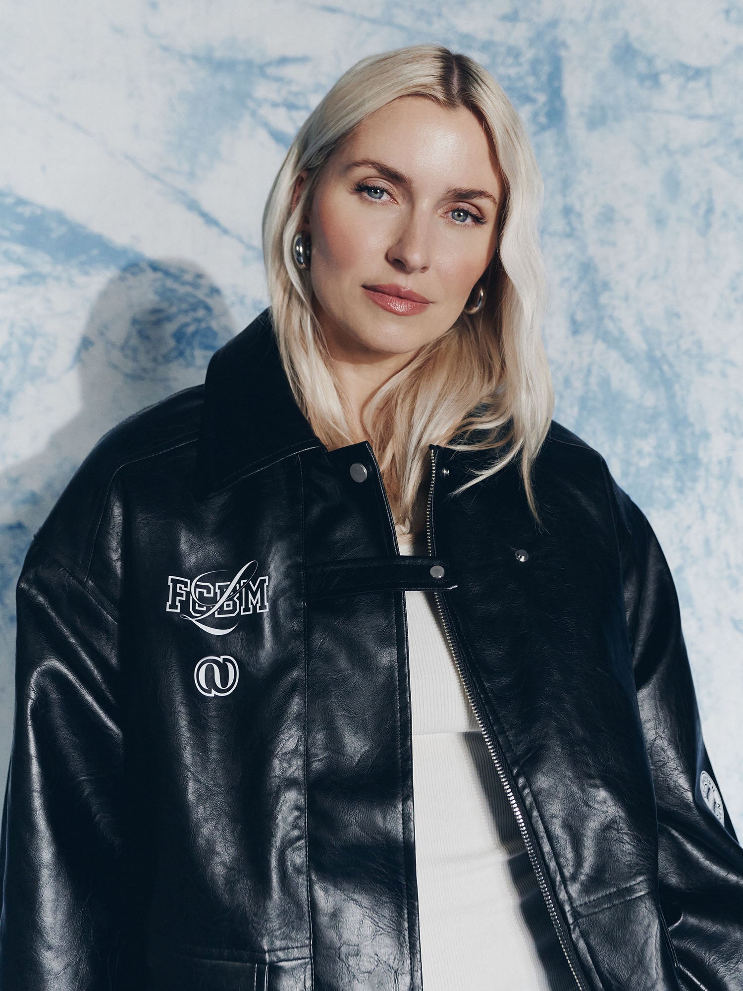 Eine Frau mit langen blonden Haaren trägt die LeGer by Lena Gercke Elisa Jacket über einem weißen Oberteil und posiert selbstbewusst vor einem blau-weiß strukturierten Hintergrund.