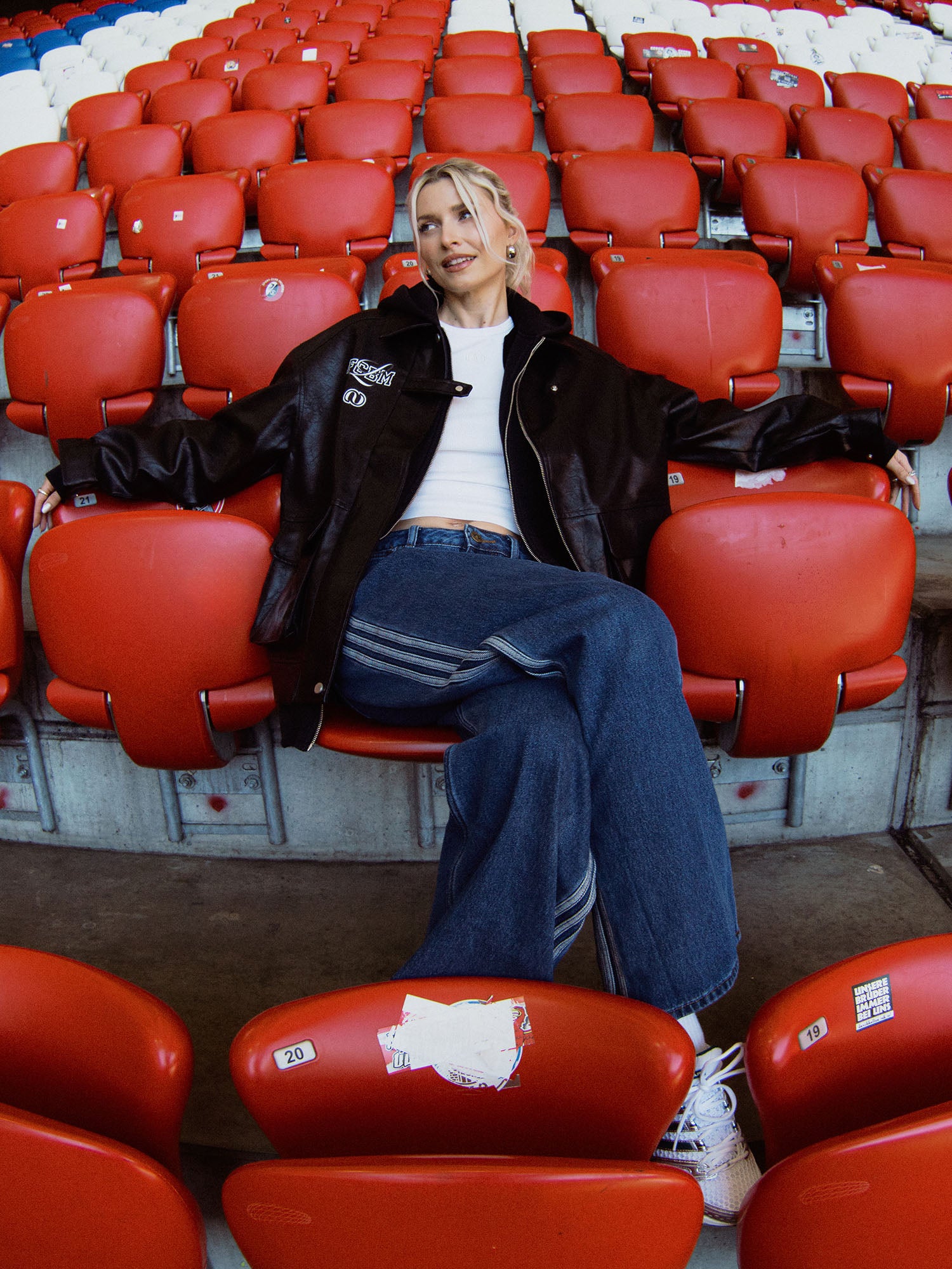 Mit der LeGer by Lena Gercke Elisa Jacket, einem weißen Crop-Top, einer weit geschnittenen Jeans und Turnschuhen entspannt sich eine Person mit ausgestreckten Armen auf den roten Stadionsitzen bei einem Spiel der FC Bayern Frauen.