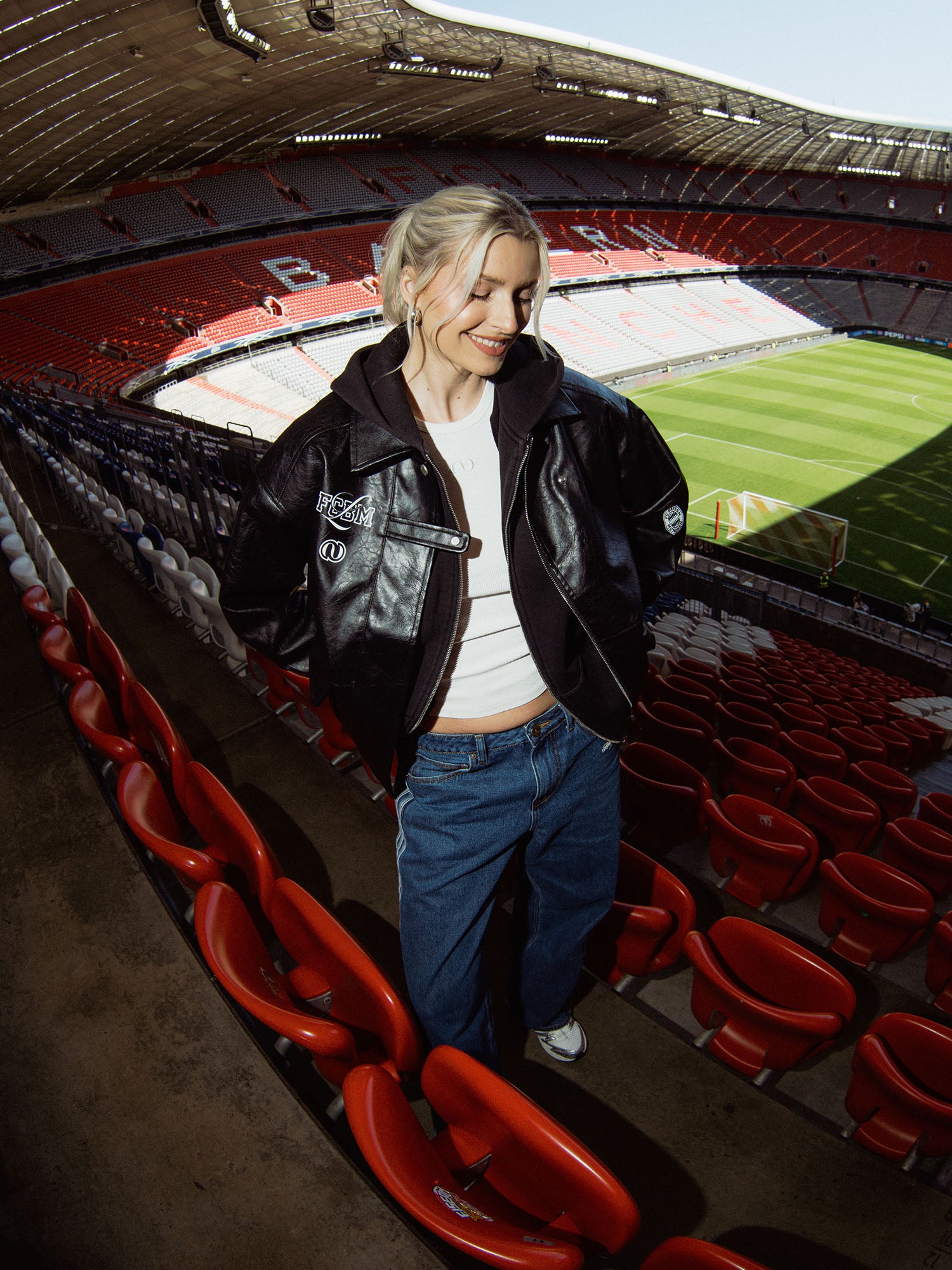 Eine lächelnde blonde Frau steht im leeren Stadion der FC Bayern Frauen. Sie trägt eine schwarze LeGer by Lena Gercke Elisa Jacke über einem weißen Hemd und einer blauen Jeans, mit dem grünen Spielfeld und den roten Sitzen im Hintergrund.