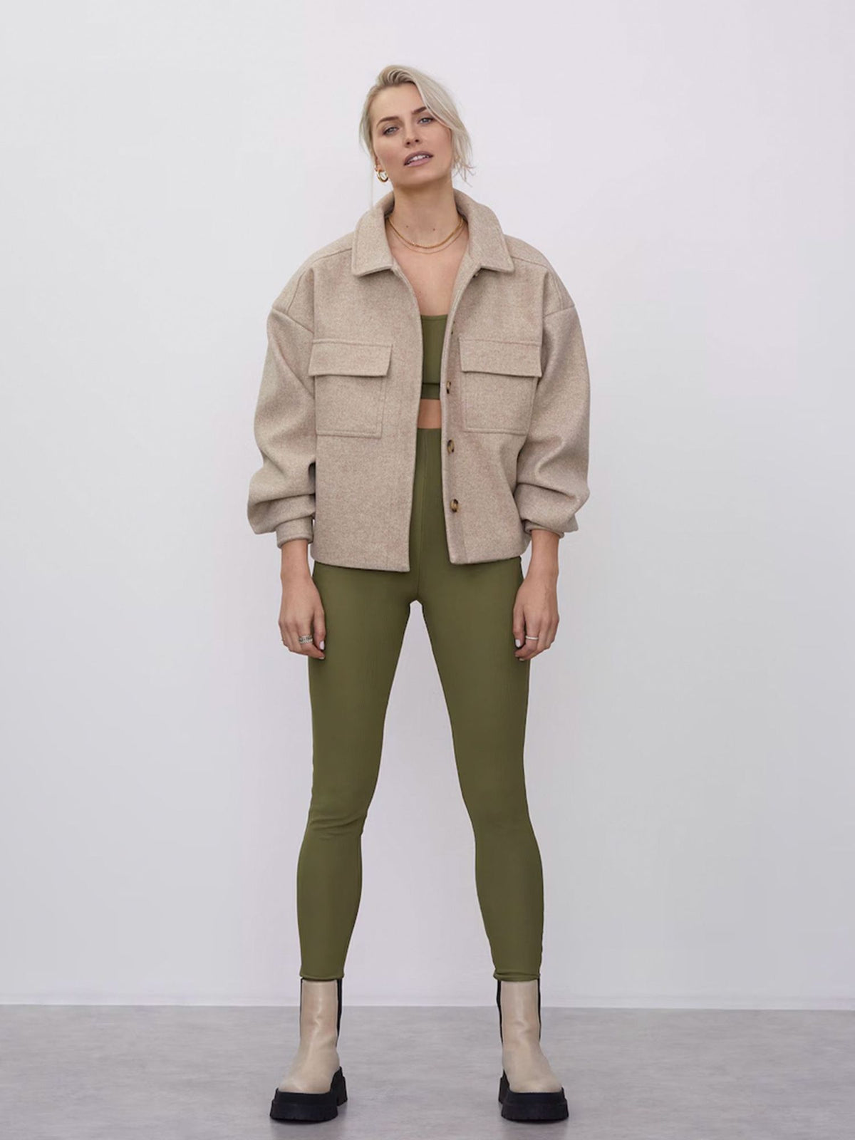 Eine Person steht vor einem schlichten Hintergrund und trägt die Jacke „Eva“ von LeGer by Lena Gercke in Beige mit Brusttaschen, dazu olivgrüne Leggings, ein passendes Oberteil und hellbraune Stiefel. Ihr helles Haar ist zu einem lockeren Pferdeschwanz zusammengebunden, und ihr Gesichtsausdruck ist neutral.