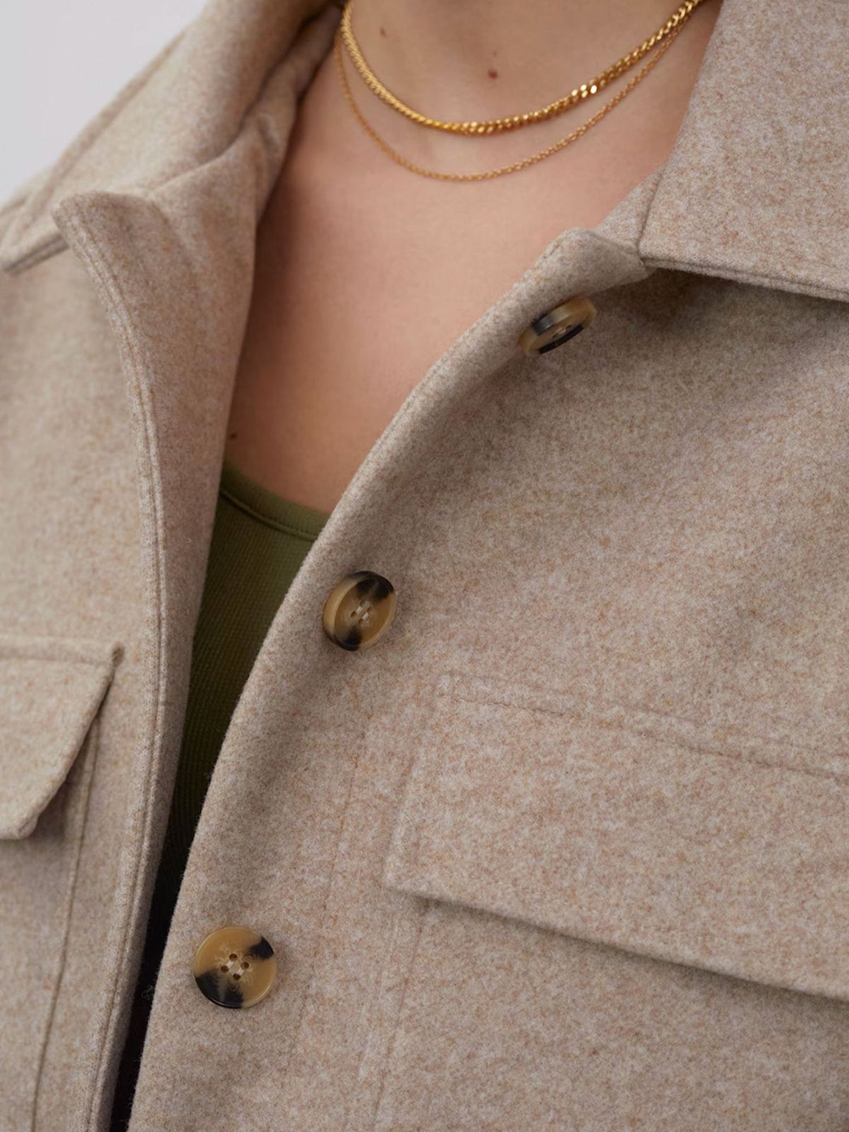 Eine Nahaufnahme einer Person, die die Jacke „Eva“ von LeGer by Lena Gercke in Hellbeige trägt – mit großen Knöpfen und Brusttaschen –, kombiniert mit einem grünen Oberteil und zwei dünnen goldenen Halsketten als Accessoires.