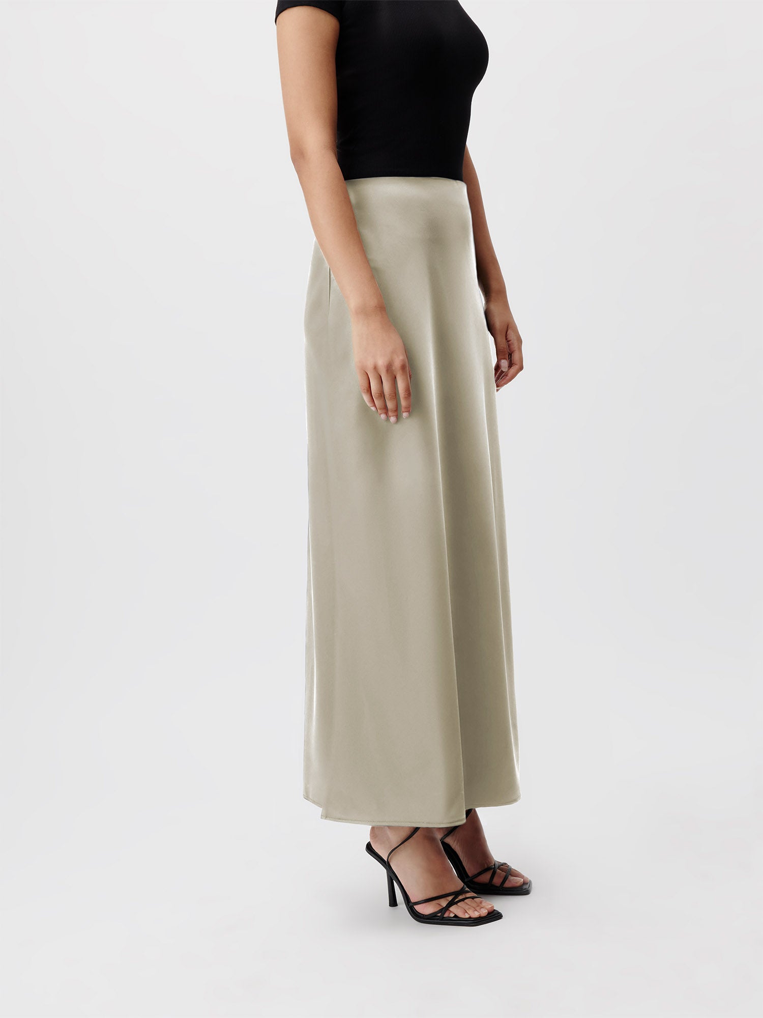 Eine Person steht vor einem weißen Hintergrund und trägt ein schwarzes Kurzarmoberteil, den langen seidigen beigefarbenen Ida-Rock von LeGer by Lena Gercke und schwarze Riemchen-High-Heels. Das Gesicht der Person ist nicht sichtbar, da das Bild von den Schultern abwärts abgeschnitten ist, was die elegante Passform des Outfits hervorhebt.