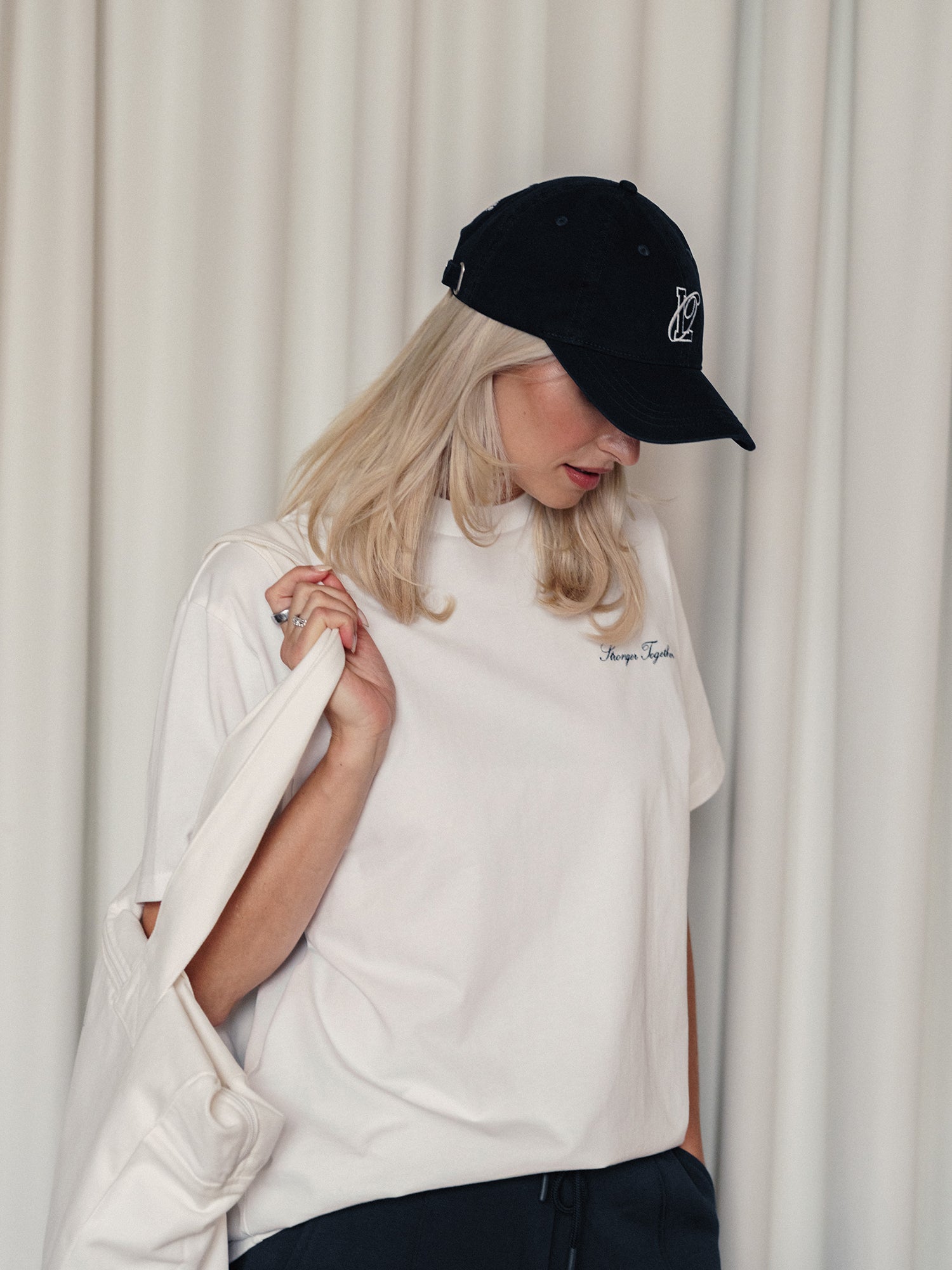 Eine Frau mit langen blonden Haaren trägt das LeGer x Oh April Amara T-Shirt Stronger von LeGer by Lena Gercke, eine schwarze Baseballkappe, eine marineblaue Hose und trägt eine weiße Tragetasche, während sie vor weißen Vorhängen steht und nach links unten schaut.