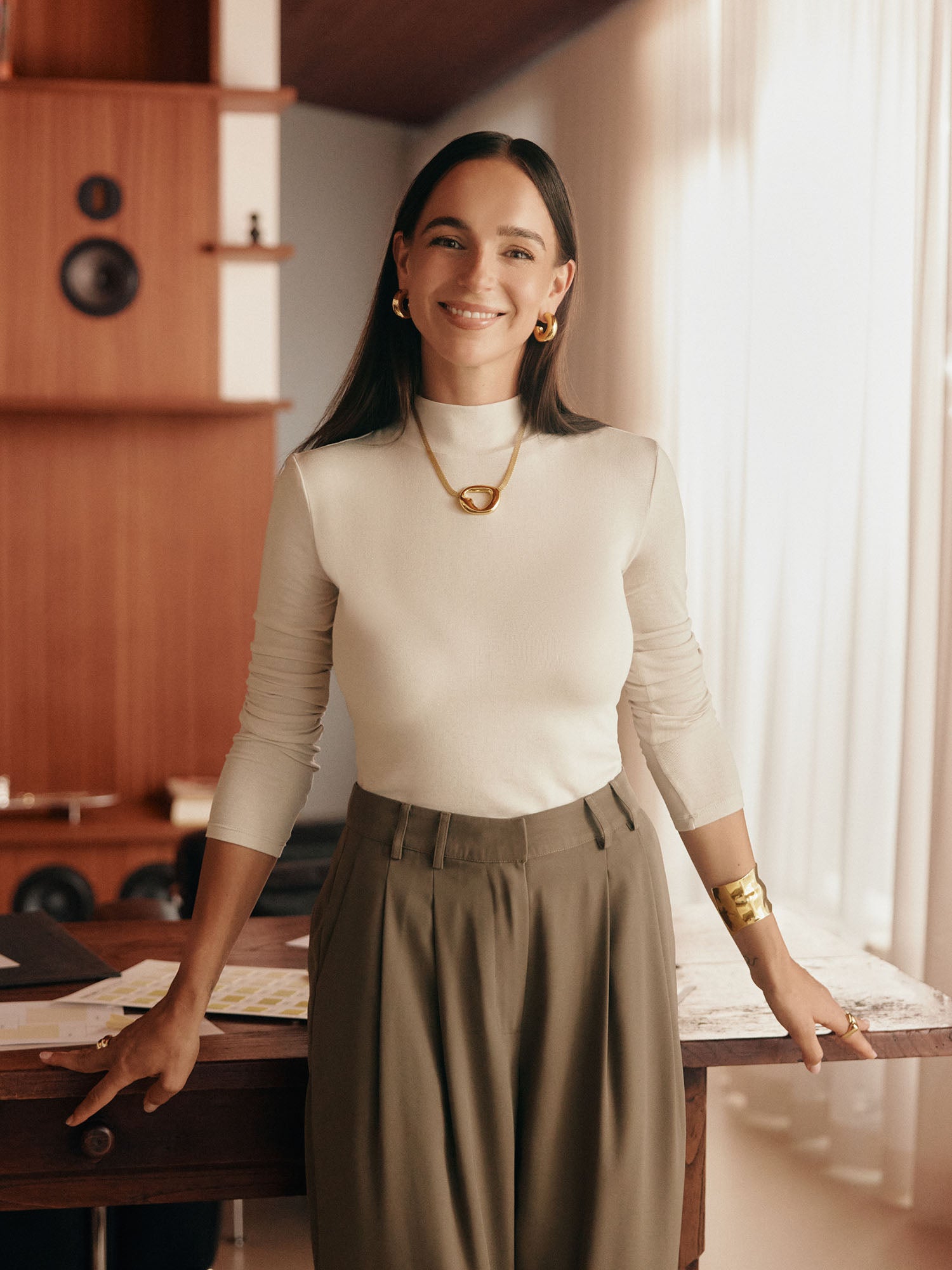 Eine Frau mit langen dunklen Haaren (Größe 36, 1,72m) lächelt an einem Schreibtisch in einem warm beleuchteten Raum. Sie trägt die LeGer x Purelei - Cara Pants von LeGer by Lena Gercke und einen weißen Rollkragenpullover. Im Hintergrund stehen Holzregale und Lautsprecher.