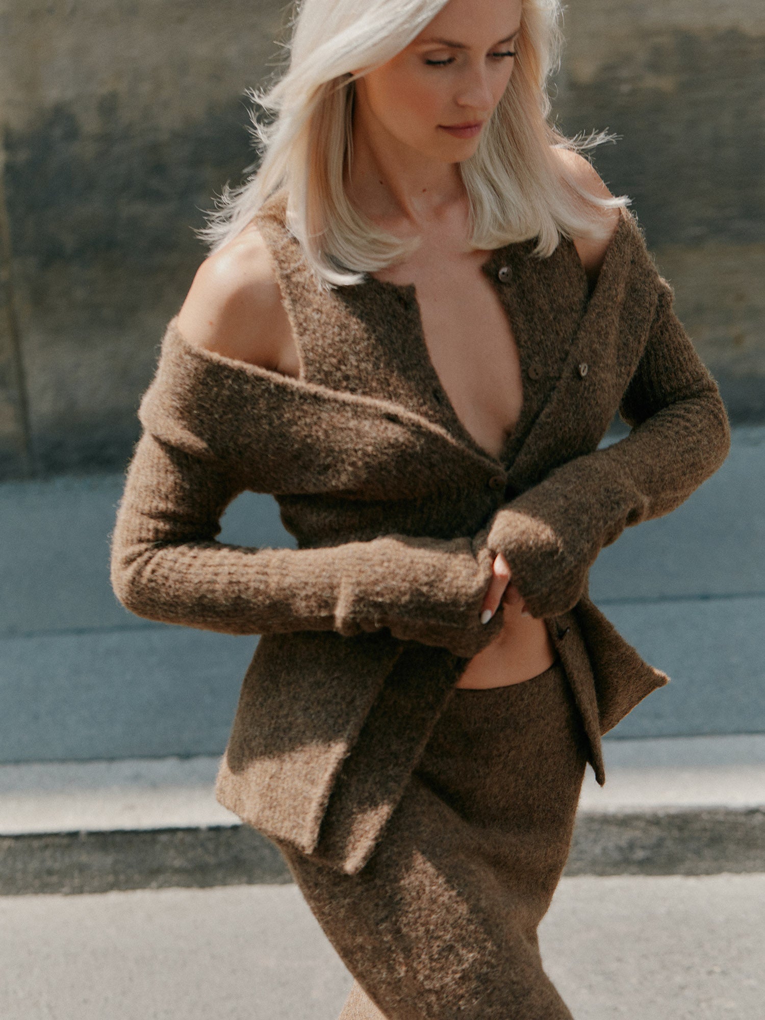 Eine Frau mit platinblondem Haar trägt das LeGer by Lena Gercke Frederike Top in Form einer braunen, strukturierten, schulterfreien Strickjacke mit passendem Rock, während sie an einem sonnigen Tag im Freien spazieren geht.