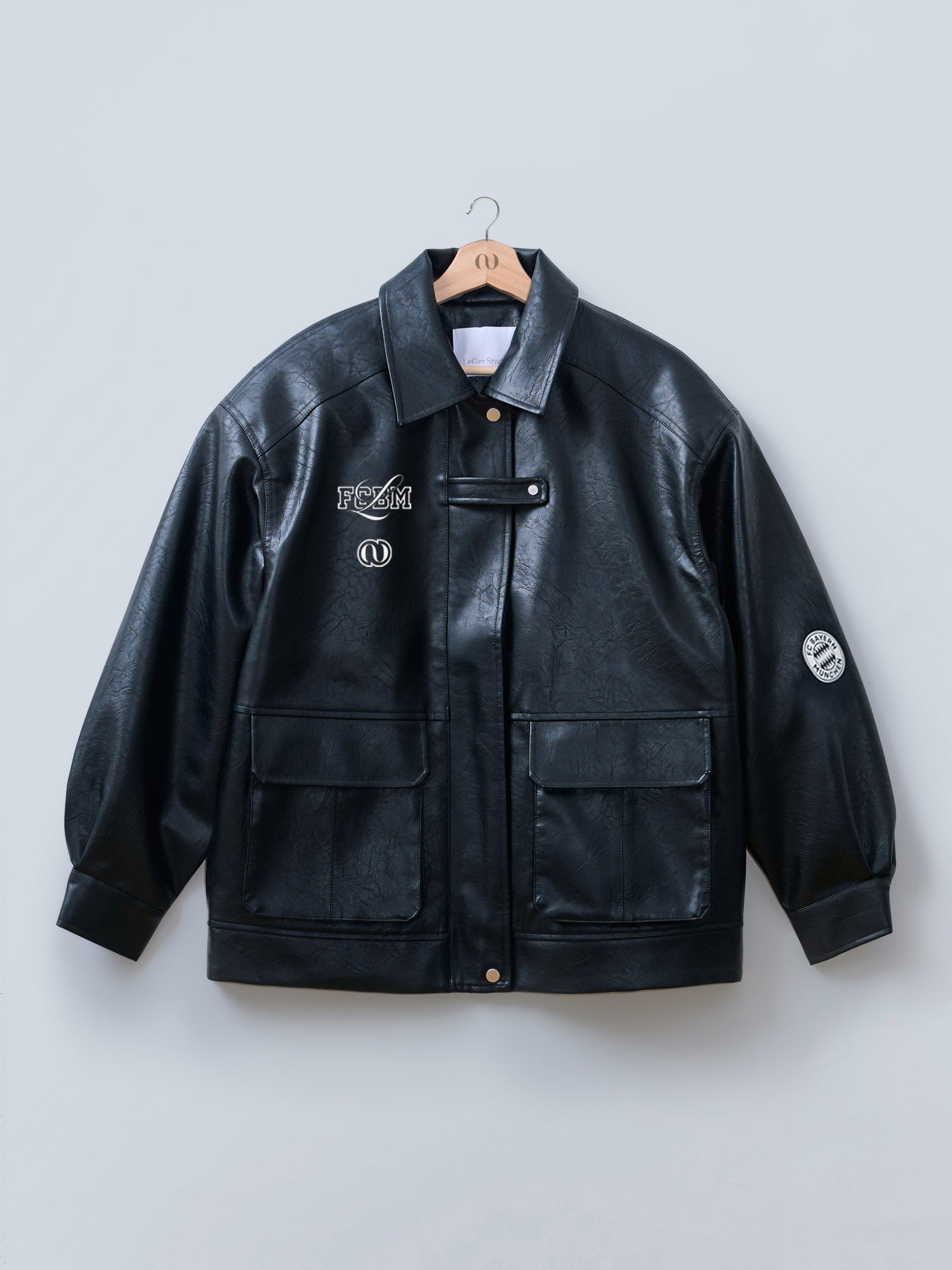 Die Elisa Jacket von LeGer by Lena Gercke ist eine schwarze Lederjacke mit zwei großen Vordertaschen, einem Kragen, Druckknöpfen, gestickten Aufnähern auf der linken Brust und einem FC Bayern Frauen-Aufnäher auf dem Ärmel. Abgebildet auf einem Holzbügel vor einem hellen Hintergrund.