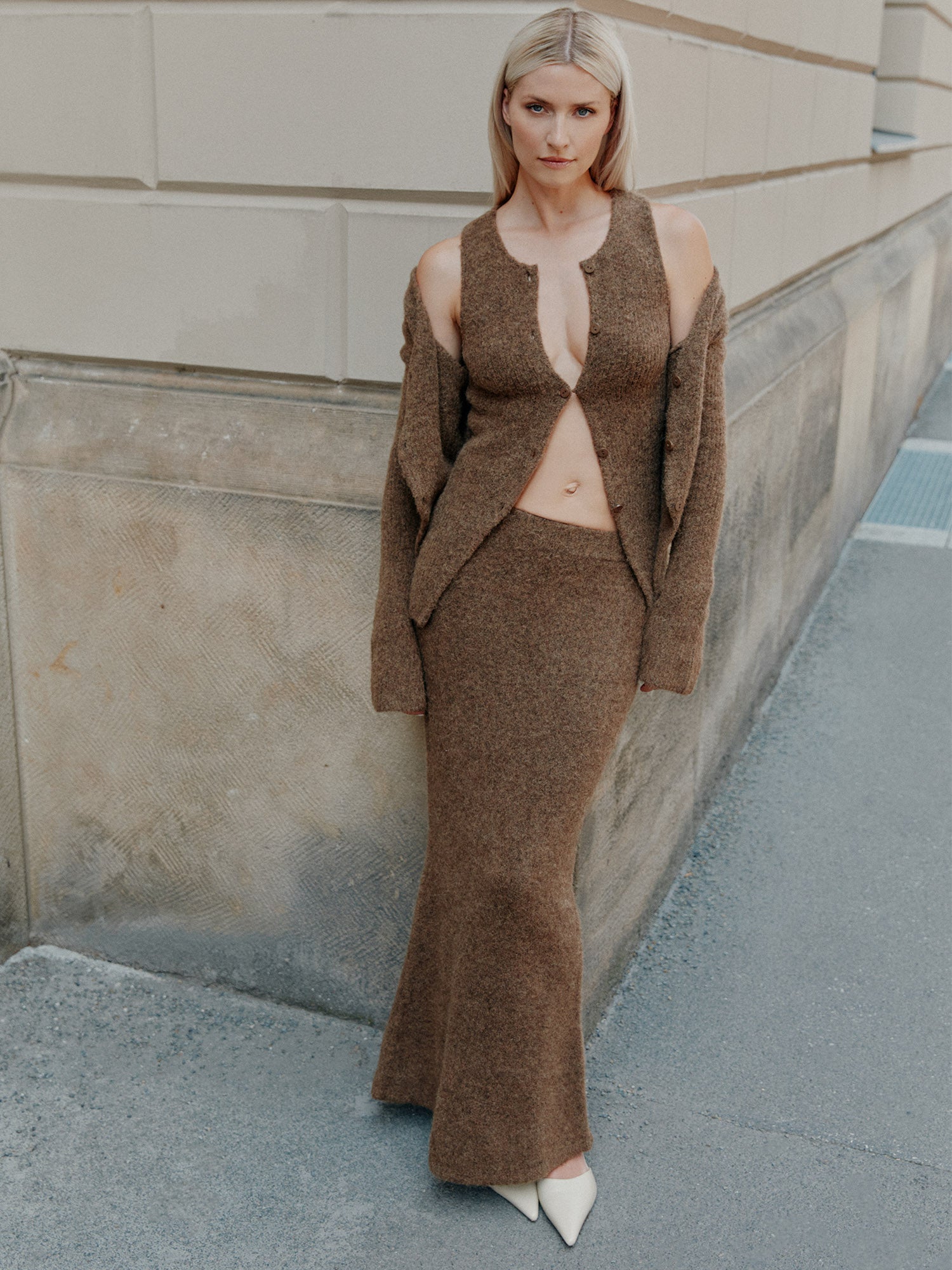 Eine Frau mit glattem blondem Haar steht draußen an einer beigen Steinmauer. Sie trägt das Frederike Top von LeGer by Lena Gercke - ein schmal geschnittenes braunes Strickteil mit tiefem Ausschnitt und langem Rock - und dazu weiße spitze Schuhe.