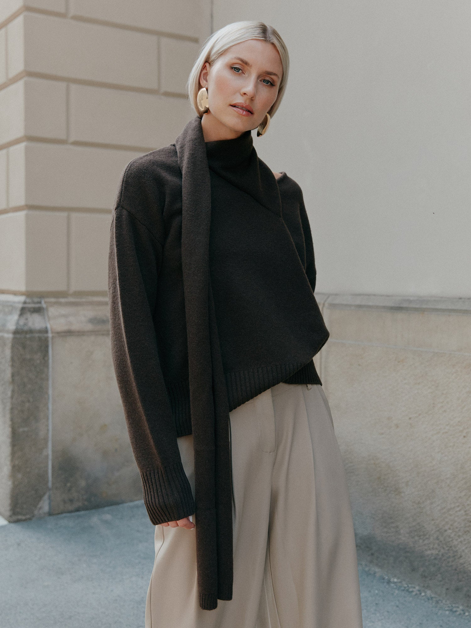 Eine Frau mit kurzen blonden Haaren trägt den Helga-Pullover von LeGer by Lena Gercke - einen locker sitzenden dunkelbraunen Pullover mit einem schalähnlichen Detail - und dazu eine weite beigefarbene Hose. Sie steht im Freien an einem hellen Gebäude und schaut neutral in die Kamera.