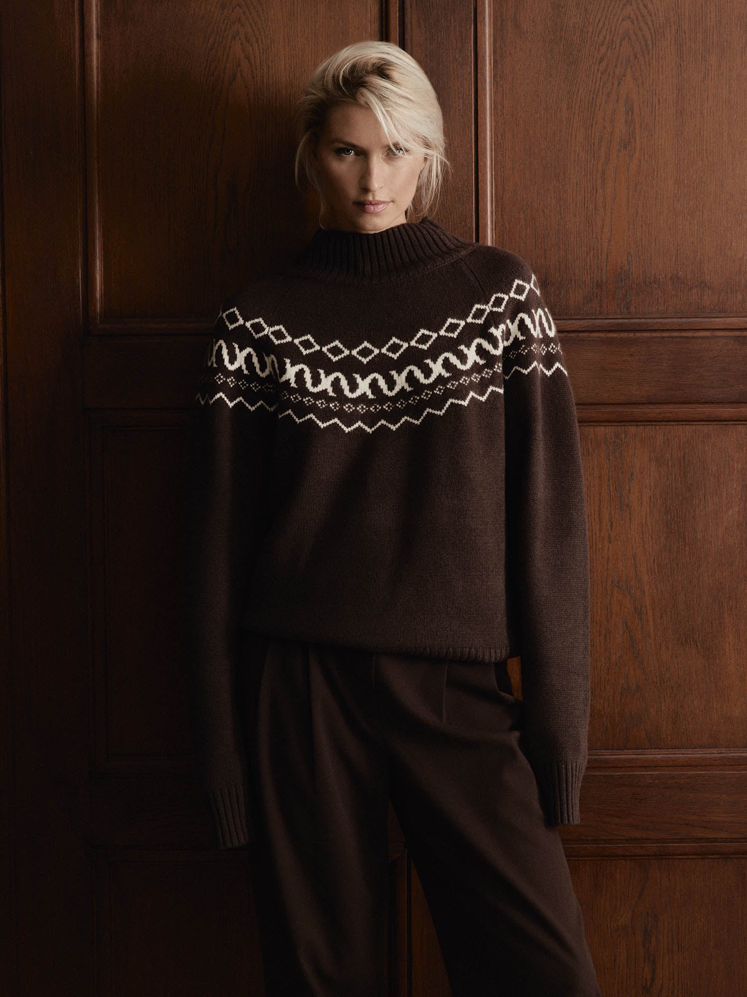 Vor einer holzgetäfelten Wand trägt eine Person mit kurzen blonden Haaren den LeGer by Lena Gercke Hellen Pullover in Dunkelbraun mit Rollkragen und weißem geometrischen Brustmuster, gepaart mit einer passenden Hose und einem neutralen Gesichtsausdruck.