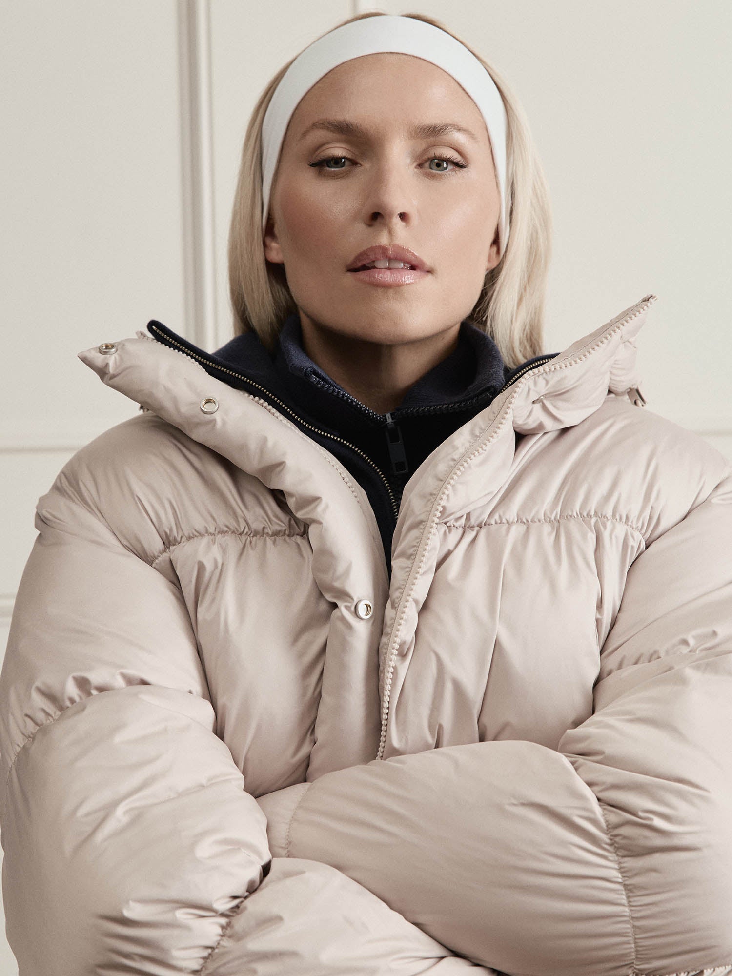 Eine Frau mit blonden Haaren trägt die Viveka Jacket von LeGer by Lena Gercke in Hellbeige (Größe 36) und ein weißes Stirnband. Sie steht mit verschränkten Armen in einem Haus und schaut selbstbewusst in die Kamera.