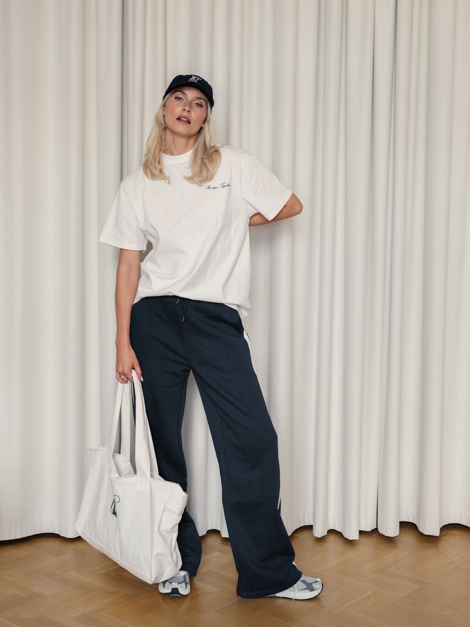Eine Person, Größe 38, steht in einem Haus und trägt eine schwarze Kappe, ein weißes T-Shirt, LeGer x Oh April - Joan Jogger Tall by LeGer by Lena Gercke in Schwarz und Turnschuhe, in der Hand hält sie eine große weiße Tragetasche mit hellen Vorhängen dahinter.